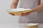 Person in a grey apron holding a large gold-finished ceramic bowl with a unique wavy white interior, a smaller matching bowl rests on the white table