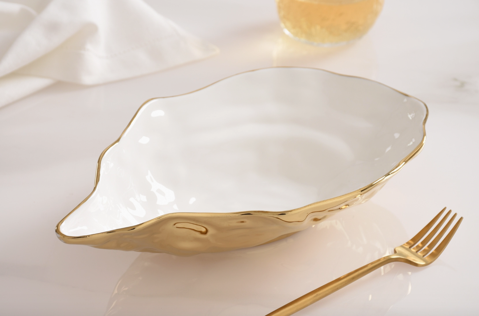 Ceramic wavy-edged white serving bowl with hammered gold exterior and matching gold fork on a white marble table next to a blurred drink and white napkin