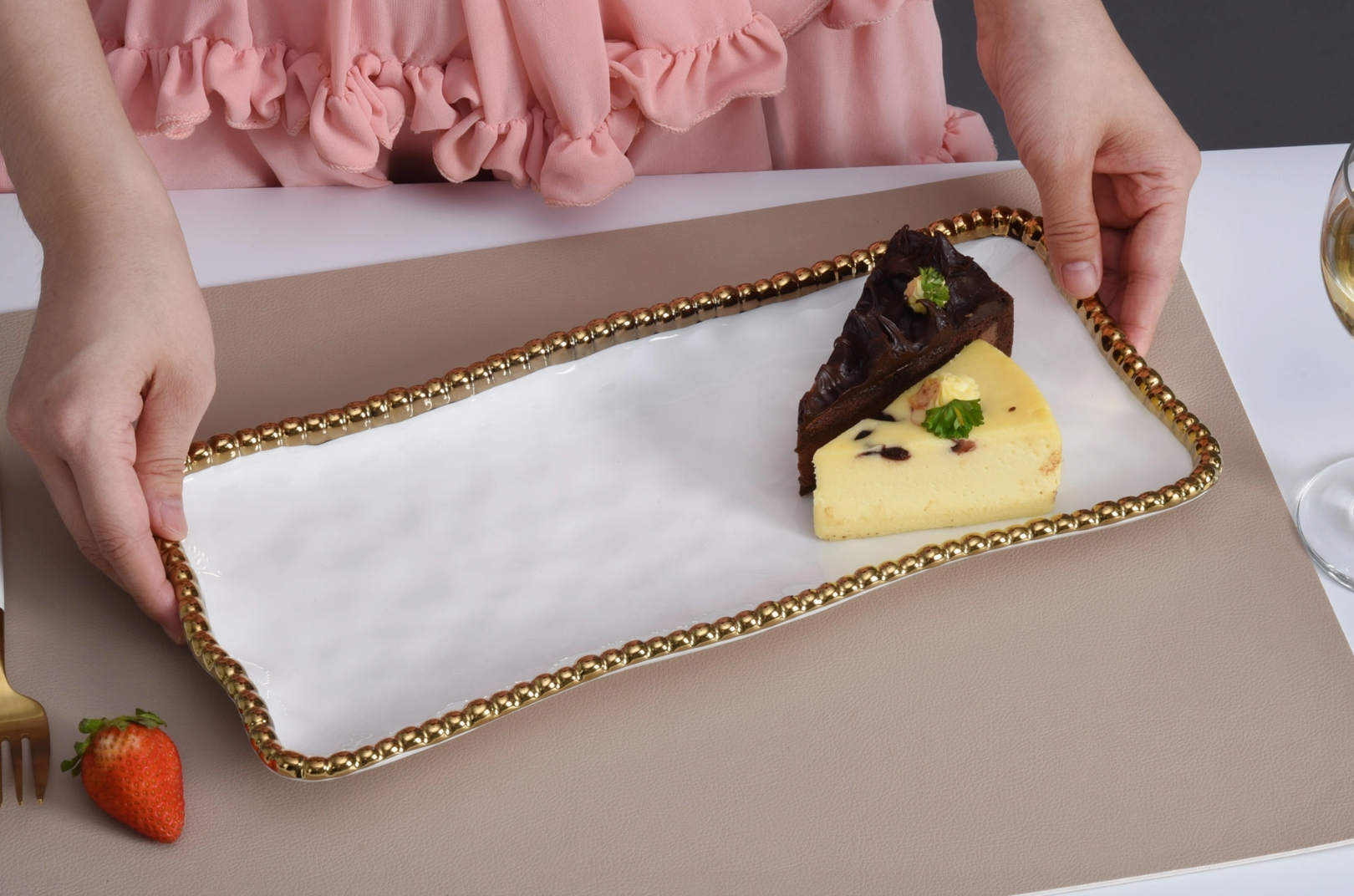 A person in a pink ruffled top presents a white rectangular serving tray with a gold beaded edge holding a dark chocolate cake slice and a yellow cheesecake slice with berry flecks and a green garnish, beside a fresh red strawberry and gold fork