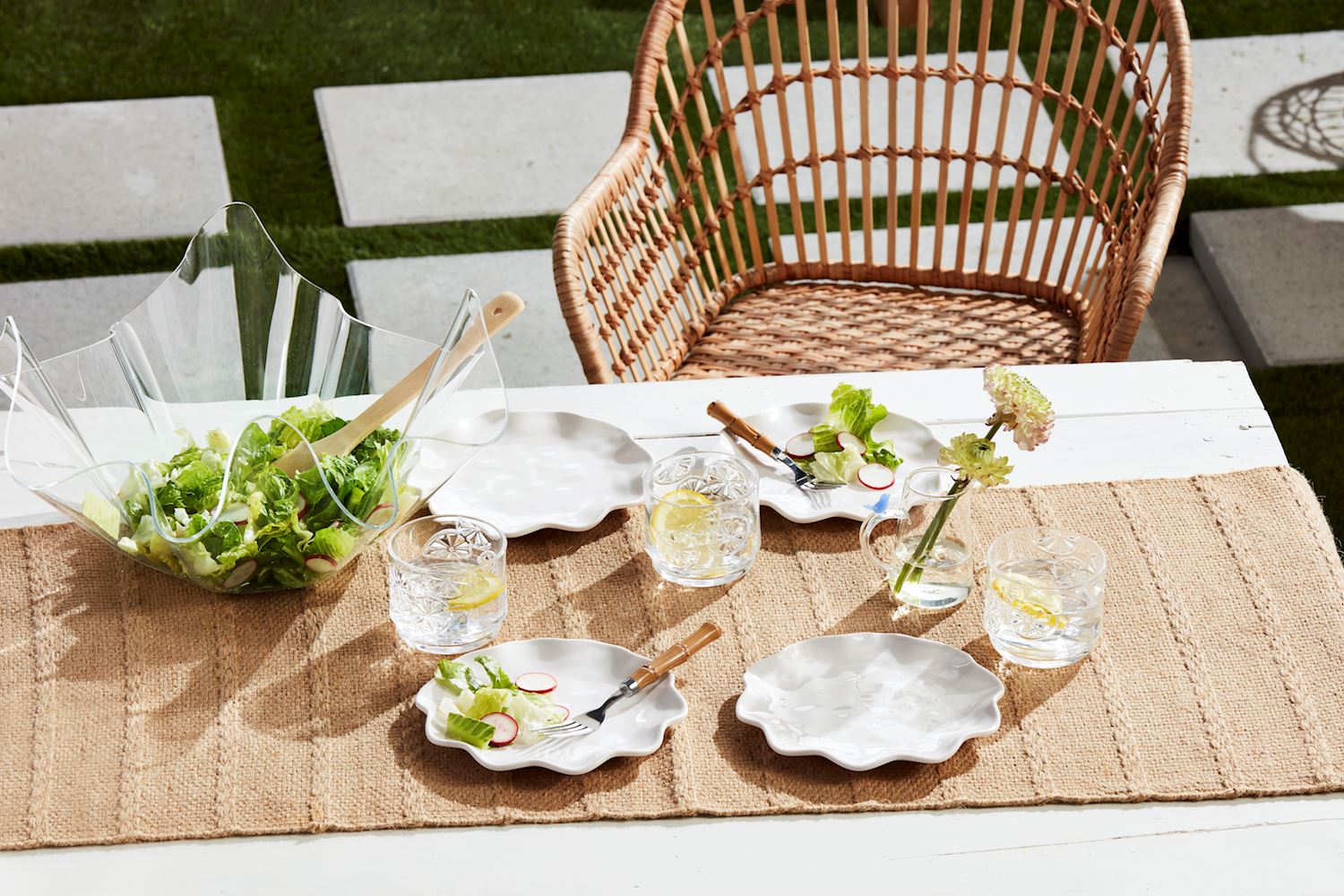 An inviting outdoor dining table setting features a natural woven table runner, white scalloped plates, a distinctive clear wavy salad bowl with fresh greens and radishes, patterned glasses with lemon slices, and a subtle pink dahlia in a small vase, all positioned beside a rustic wicker chair on a patio with green grass