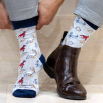 A person's hands pulling on light gray novelty socks featuring red crawfish, white pelicans, and blue crabs, next to a dark brown leather ankle boot and gray trousers on a wooden floor