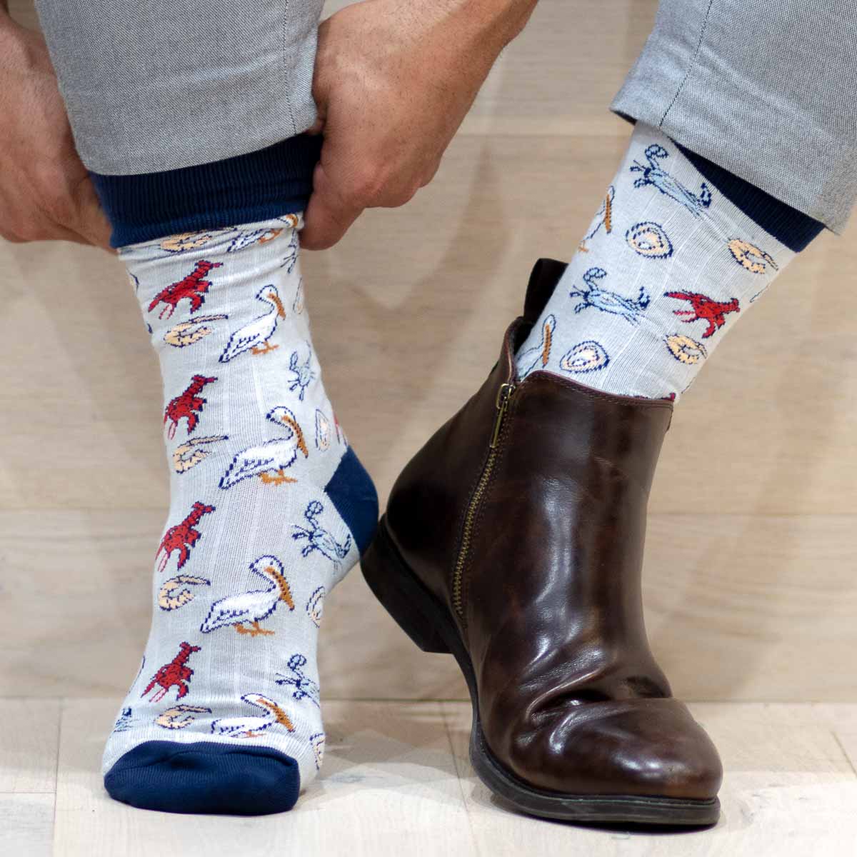 A person's hands pulling on light gray novelty socks featuring red crawfish, white pelicans, and blue crabs, next to a dark brown leather ankle boot and gray trousers on a wooden floor
