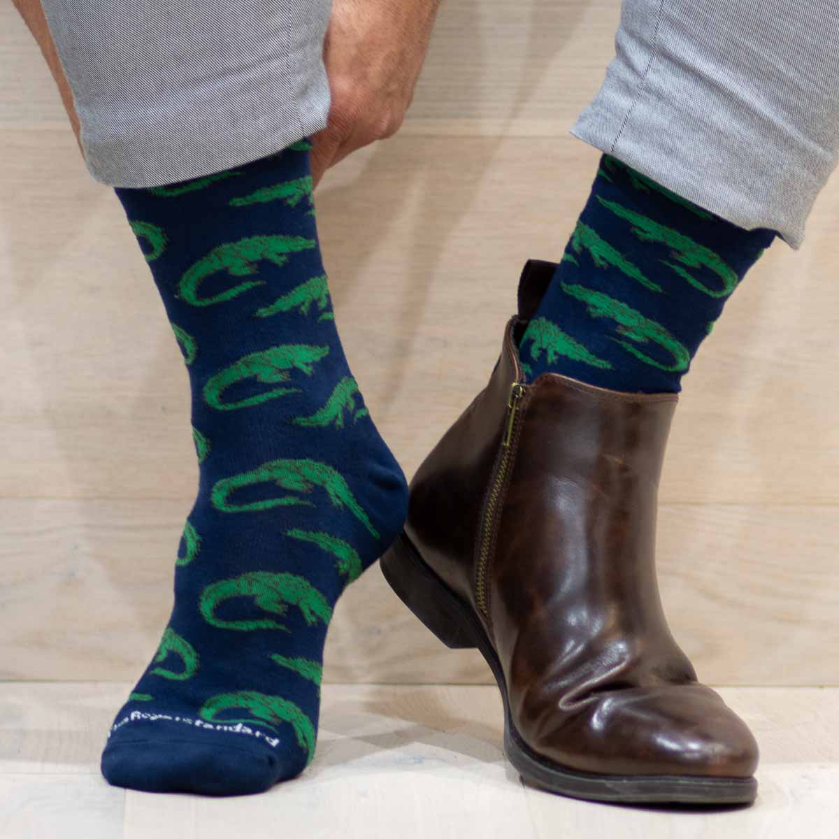 Photography of a person's lower legs on a light wood floor, one foot showcasing a navy blue Royal Standard sock with a vibrant green alligator pattern and the other foot in a dark brown leather ankle boot featuring a brass side zipper