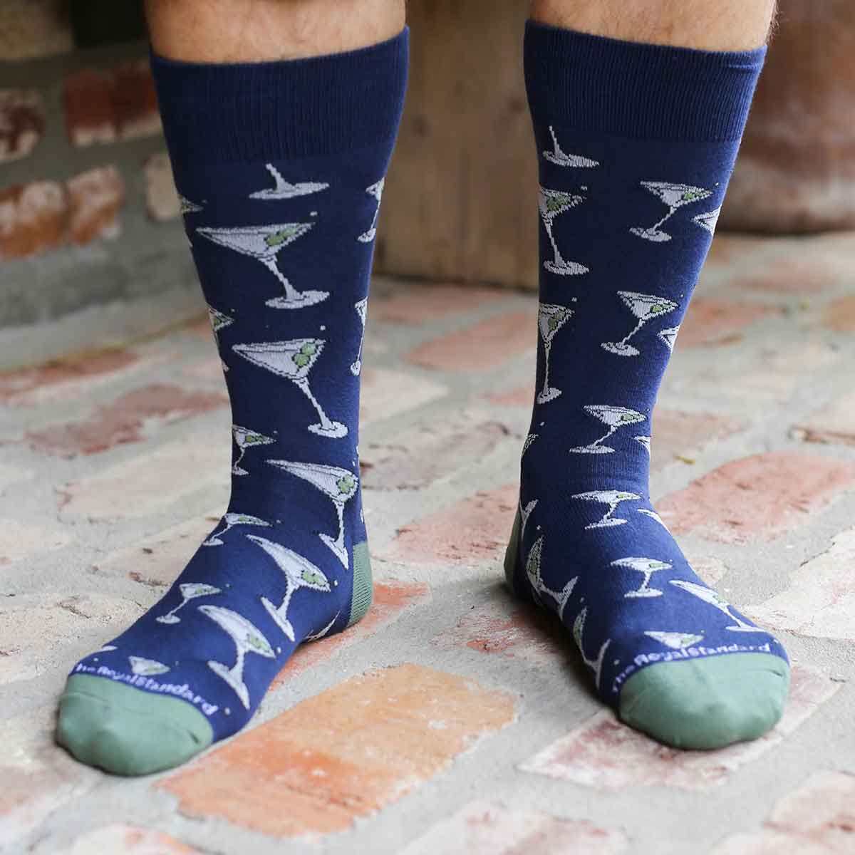 Photograph by The Royal Standard of a person wearing navy blue socks with a repeating white martini glass and green olive pattern while standing on a weathered brick patio showcasing sage green toes and heels