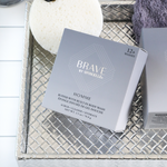 Brave by Spongellé Homme buffer with built-in body wash box featuring metallic silver 'Brave' logo and '12+ washes' on a textured silver woven tray with a white sponge and a folded purple towel