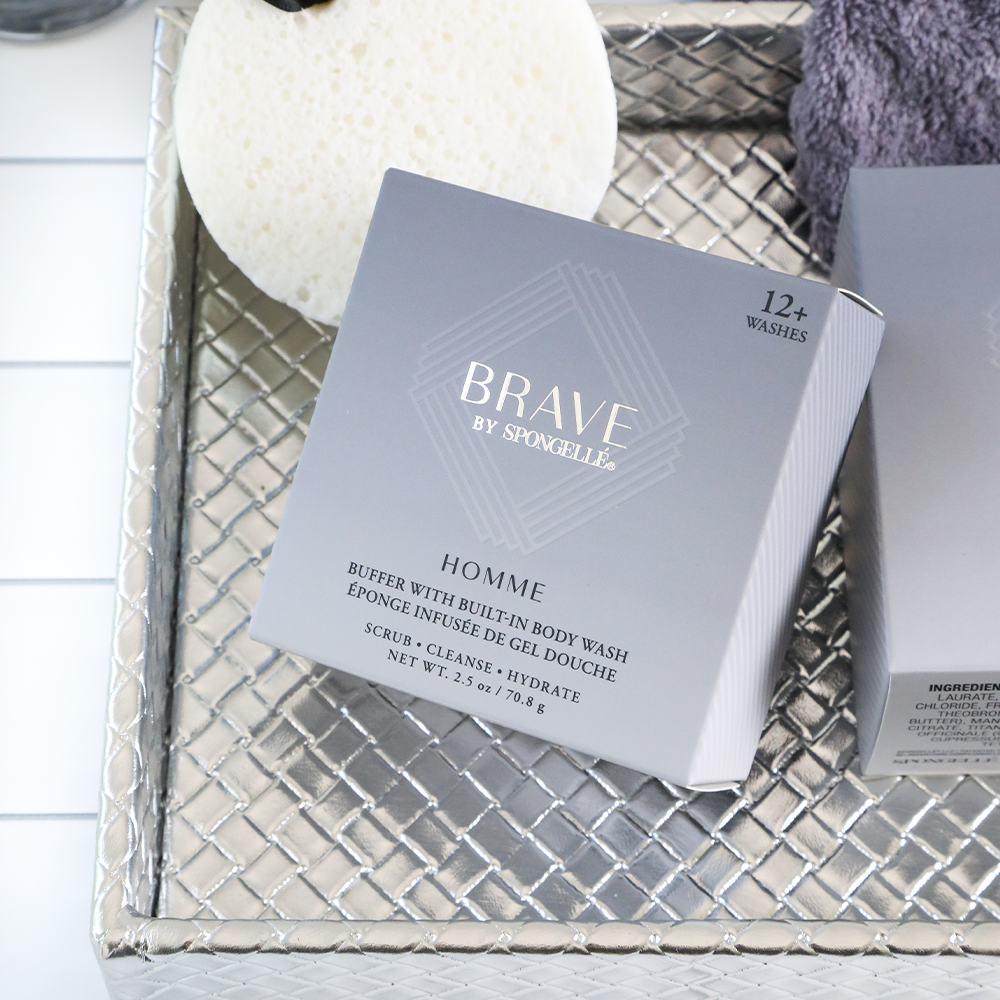 Brave by Spongellé Homme buffer with built-in body wash box featuring metallic silver 'Brave' logo and '12+ washes' on a textured silver woven tray with a white sponge and a folded purple towel