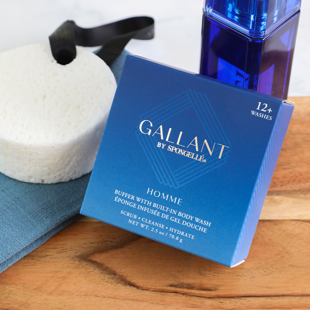 Spongellé Gallant Homme men's body care collection on a wooden surface, featuring a vibrant deep blue product box, a white round body buffer with a black ribbon, and a tall dark blue glass bottle