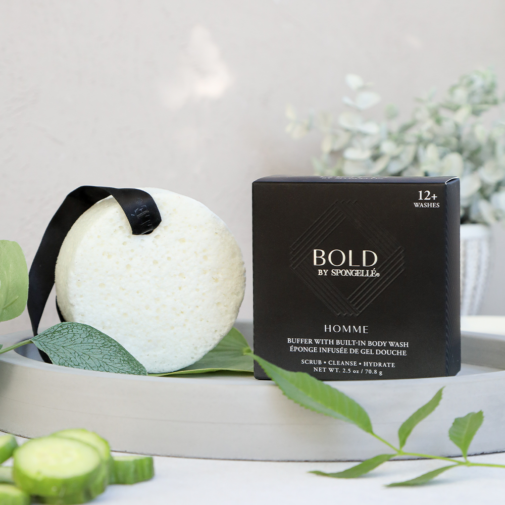 Photograph of a Bold by Spongellé Homme white circular body wash buffer with a black ribbon next to its black product box displayed on a grey concrete tray with fresh green leaves and sliced cucumbers