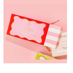 A vibrant red and pink Musee Merry Christmas gift box with gold lettering and a pink and white striped side panel sits next to a white Christmas tree shaped bath bomb on a soft pink surface