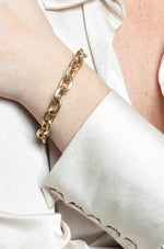 Close-up of a wrist adorned with a chunky gold-toned chain link bracelet against a cream-colored silk-like blouse featuring a distinctive scalloped cuff