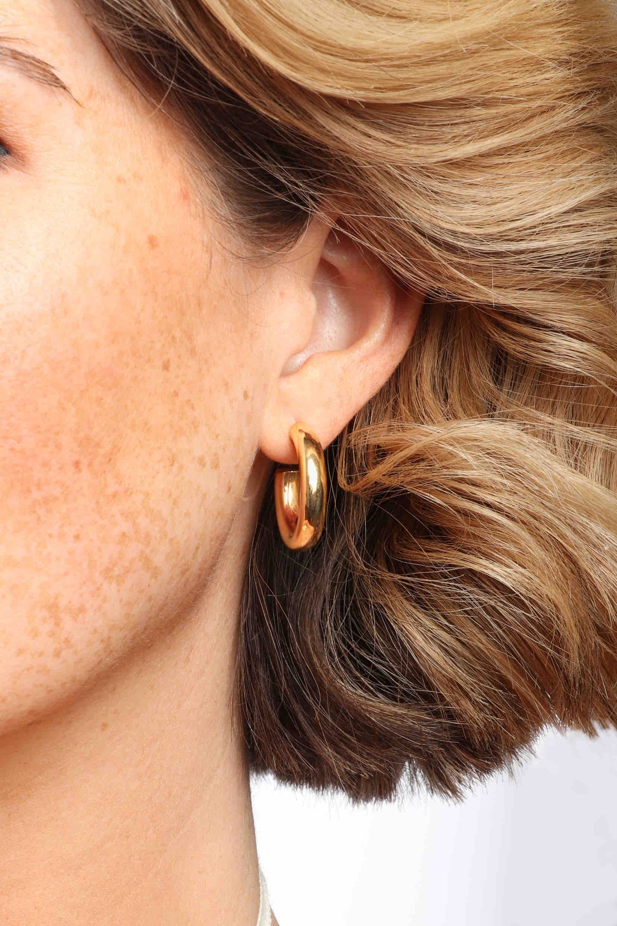 Photograph of a woman's ear and side of her face featuring a chunky gold hoop earring, blonde wavy hair, and freckled skin