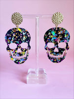A pair of black acrylic skull earrings with iridescent multicolored confetti flakes, detailed with cut-out eye sockets and teeth, hanging from textured gold-tone studs on a clear display stand against a vibrant pink backdrop