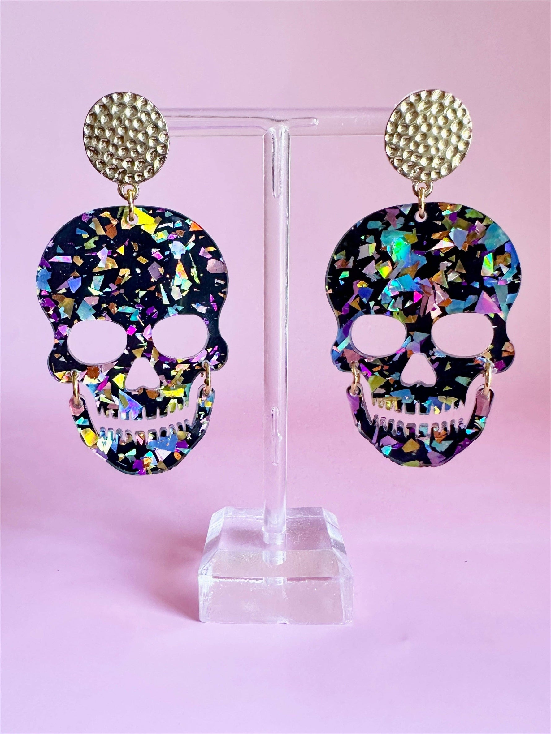 A pair of black acrylic skull earrings featuring iridescent multicolor glitter shards, cut-out eye sockets, and teeth, suspended from hammered gold-tone studs on a clear display stand against a light pink background