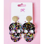 Acrylic earrings by S&R featuring two black skull designs with iridescent confetti glitter and hammered gold stud tops, displayed on a white card against a vibrant pink background