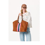 Woman wearing a brown baseball cap and white embroidered sweatshirt holding a brown suede tote bag with striped detail