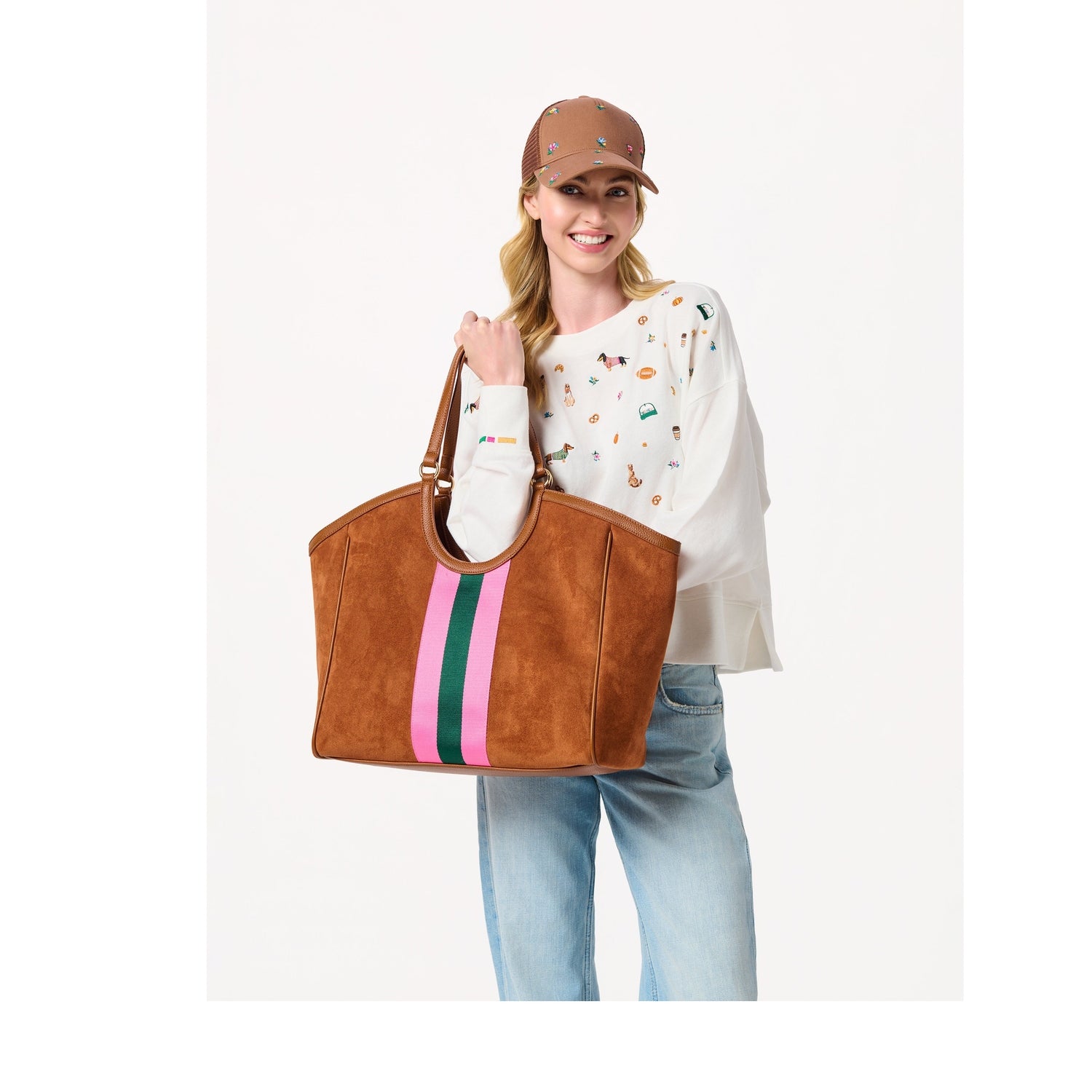 Woman wearing a brown baseball cap and white embroidered sweatshirt holding a brown suede tote bag with striped detail