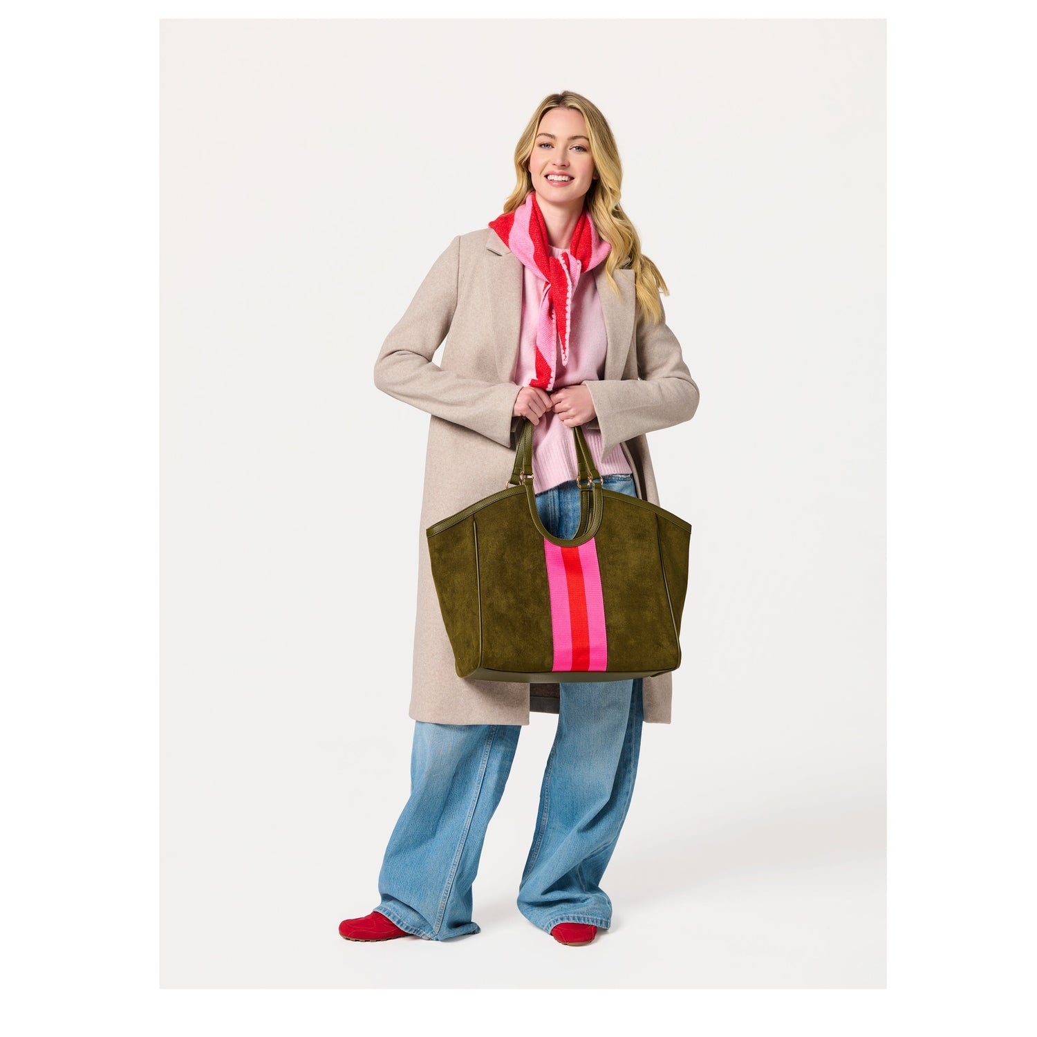 Woman wearing a beige coat, pink sweater, and jeans holding an olive green suede tote bag with a pink and red stripe
