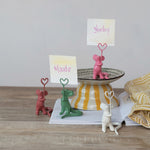 Ceramic playful mouse place card holders in pink, green, reddish-brown, and white, each holding a heart-shaped wire clip with names like Maude and Stanley, arranged on a wooden table alongside a yellow striped pedestal bowl and an open magazine