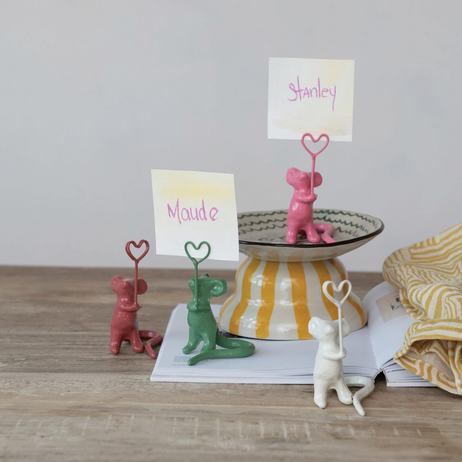 Ceramic playful mouse place card holders in pink, green, reddish-brown, and white, each holding a heart-shaped wire clip with names like Maude and Stanley, arranged on a wooden table alongside a yellow striped pedestal bowl and an open magazine