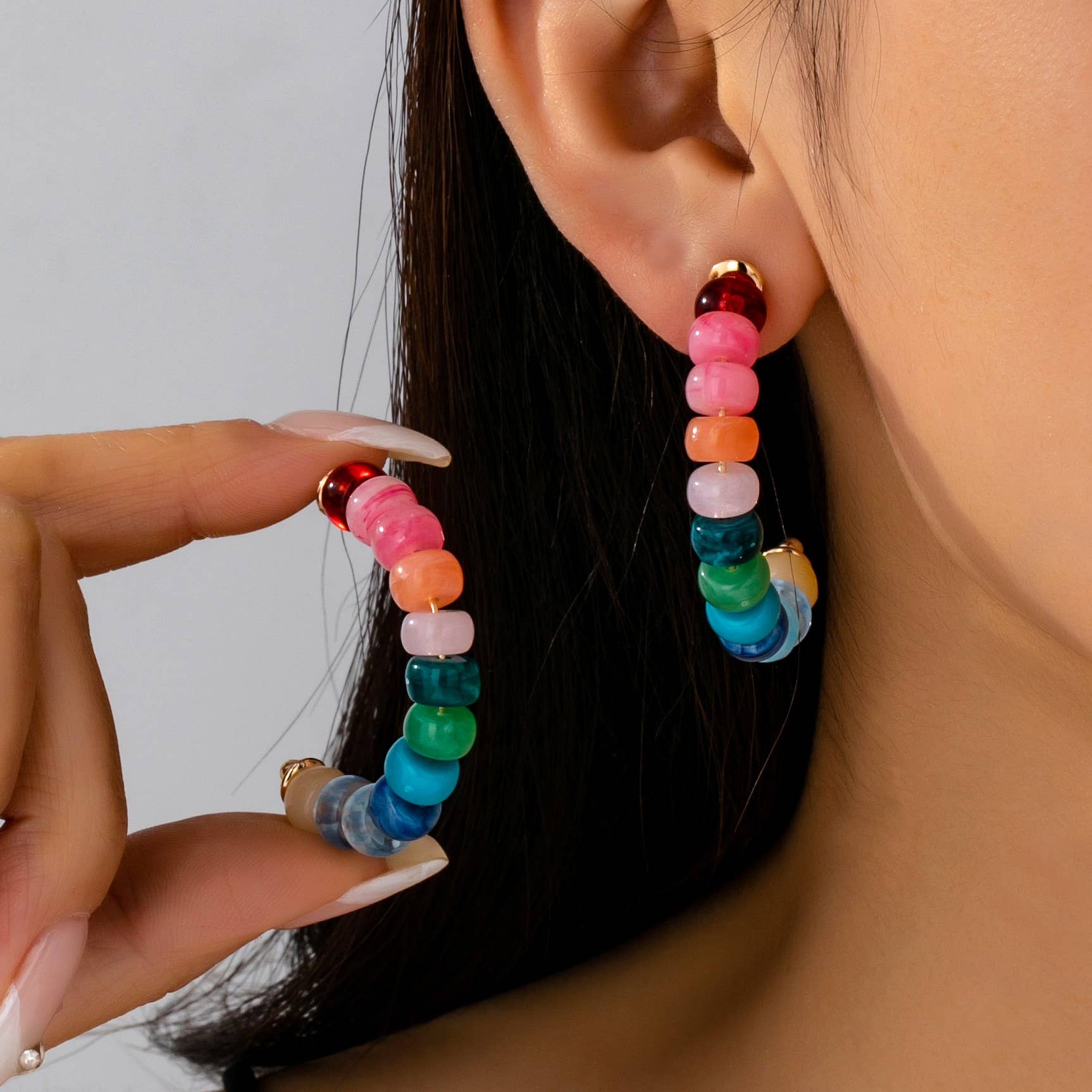 Close-up of a person's ear showcasing a dangling rainbow gradient beaded hoop earring with gold-toned findings while a hand with manicured nails holds a matching earring against a neutral backdrop