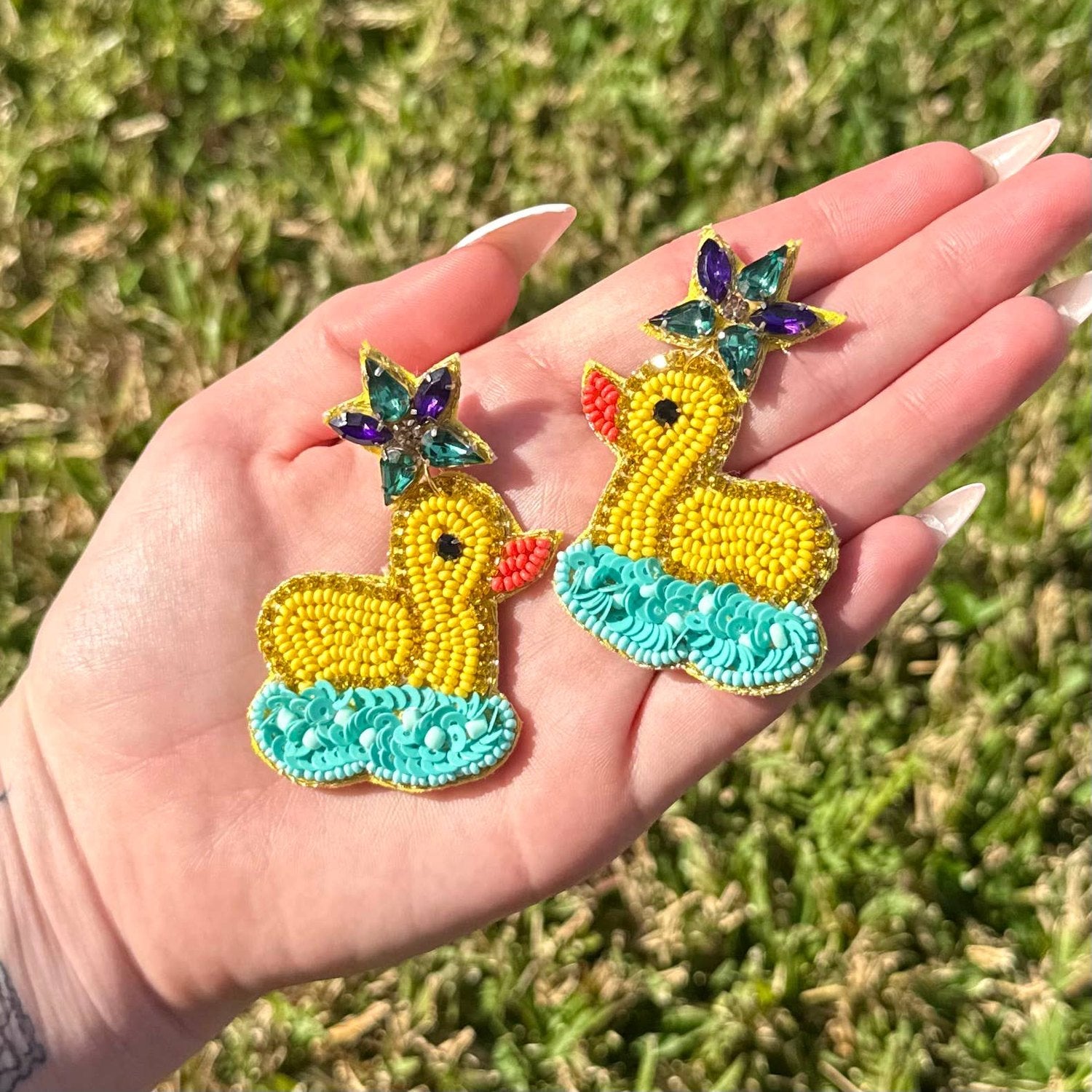 Beaded duck earrings held in a hand featuring vibrant yellow ducks with red beaks floating on teal beaded waves topped with sparkling purple and teal star-shaped jewels against a green grass background