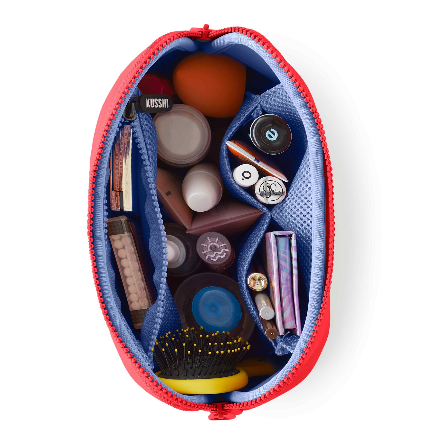 An overhead product shot of a red KUSSHI makeup bag unzipped on a white background, revealing a blue mesh interior organized with an orange beauty blender, a yellow detangling brush, and various cosmetic bottles