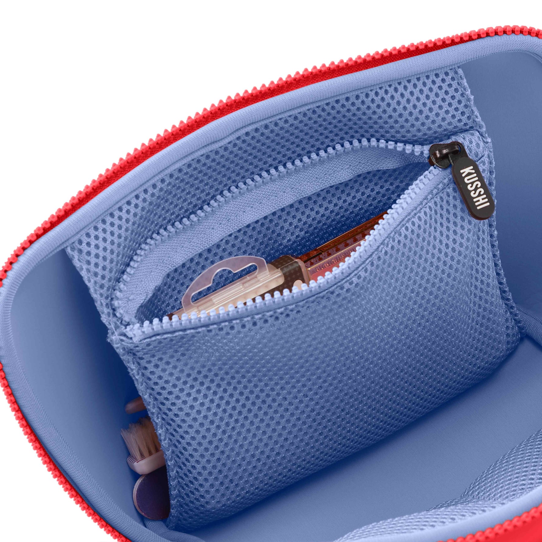 A Kusshi makeup bag interior featuring a bright red zipper, a periwinkle blue lining, a mesh zippered pocket with a KUSSHI pull, and a small brush and compact inside