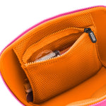 Digital photograph of an open KUSSHI makeup bag in a studio setting showcasing its vibrant pink exterior zipper, bright orange interior lining, and an orange mesh pocket with a KUSSHI branded zipper pull, holding a small brush and compact