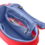 Product photo of an open KUSSHI cosmetic bag with a coral red exterior and periwinkle blue interior, revealing a mesh zipper pocket holding a pink beauty tool and featuring a branded KUSSHI zipper pull on a white studio background