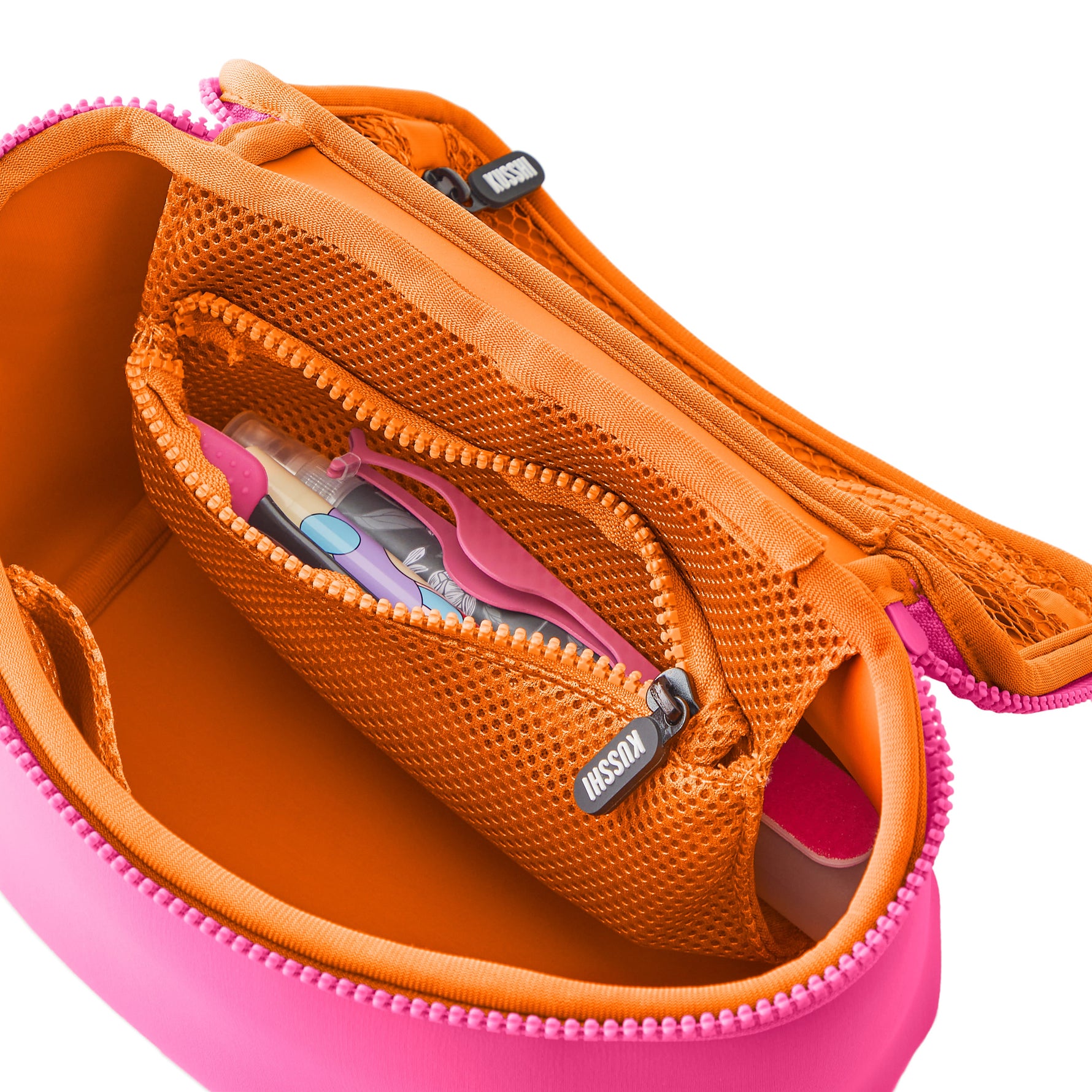 Open KUSSHI makeup organizer bag on a white background featuring a bright fuchsia pink exterior and vibrant orange interior with a mesh zippered pocket containing tweezers and a roller bottle