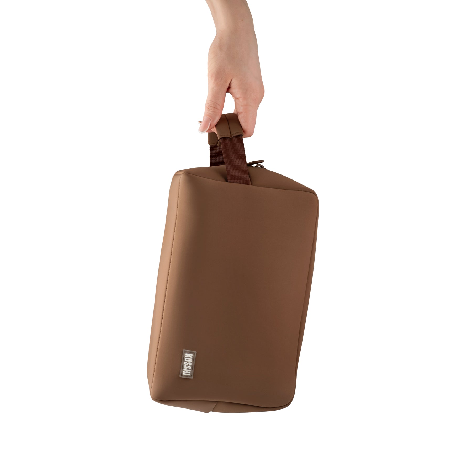 A hand holds a brown KUSSHI rectangular pouch bag featuring a silver-toned KUSSHI logo and woven strap handle on a clean white background