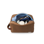 Photograph of a KUSSHI brown neoprene toiletry bag with its logo visible, open on a white background, revealing a light blue interior lining and brown zipper, filled with travel-sized Weleda toothpaste, Coach Open Road lotion, a white soap bar, and a Philips shaver