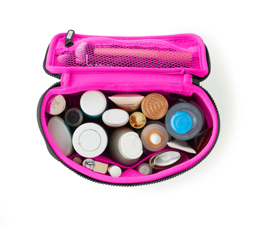 An overhead studio shot of a neon pink and black travel makeup bag unzipped, revealing various white, gold, and bronze cosmetic containers, a bottle with a bright blue cap, and makeup brushes in an internal mesh pocket