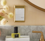 NEST New York's gold-framed wall diffuser and vibrant Grapefruit oil bottle sit on a marble countertop amidst lush white dahlia flowers and an elegant brass faucet