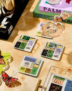 Photograph of a vibrant tabletop setting with New Orleans themed matchbox sets featuring Cafe Du Monde, Bourbon Street, and The Big Easy NOLA, a champagne glass topped with a brown butterfly, a red butterfly ornament, and decorative books for Palm Beach and Miami