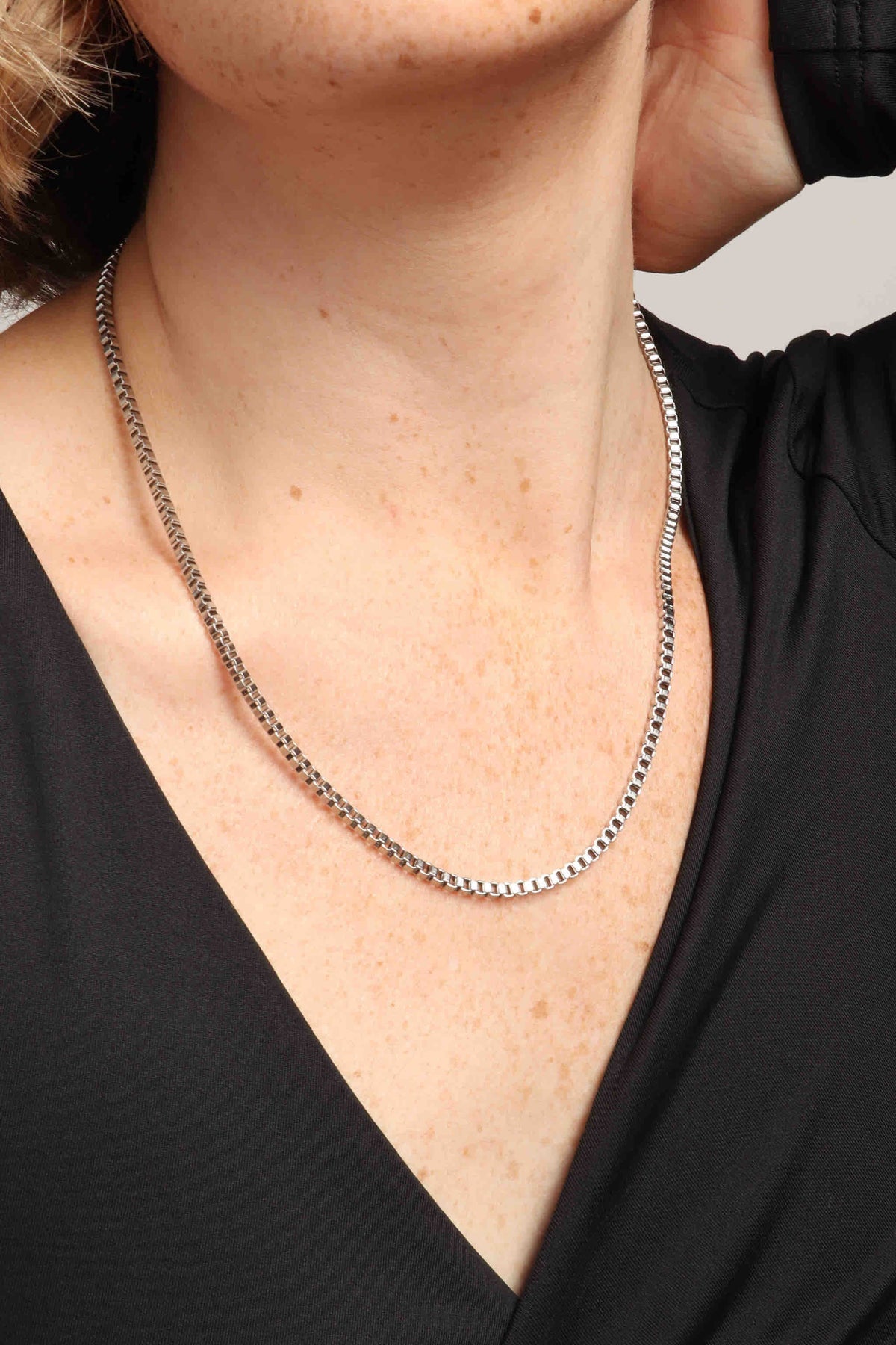 Close-up of a person's freckled neck and upper chest wearing a shiny silver box chain necklace over a black v-neck top