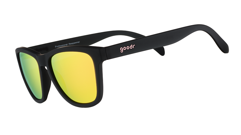 A pair of Goodr Professional Respawner sunglasses with matte black frames, vibrant yellow-green mirrored lenses, and a pink goodr logo on the temple, resting on a clear acrylic stand