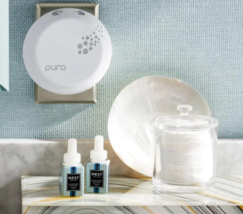 A white Pura smart diffuser with a grey dot pattern is plugged into a light blue textured wall above two Nest Ocean Mist & Sea Salt blue fragrance refills on a marble vanity with a glass jar of cotton pads and an iridescent shell-like dish