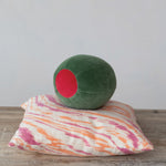 A green velvet spherical cushion with a red circle detail rests on a rectangular cushion featuring a vibrant pink and orange abstract striped pattern all displayed on a rustic wooden surface