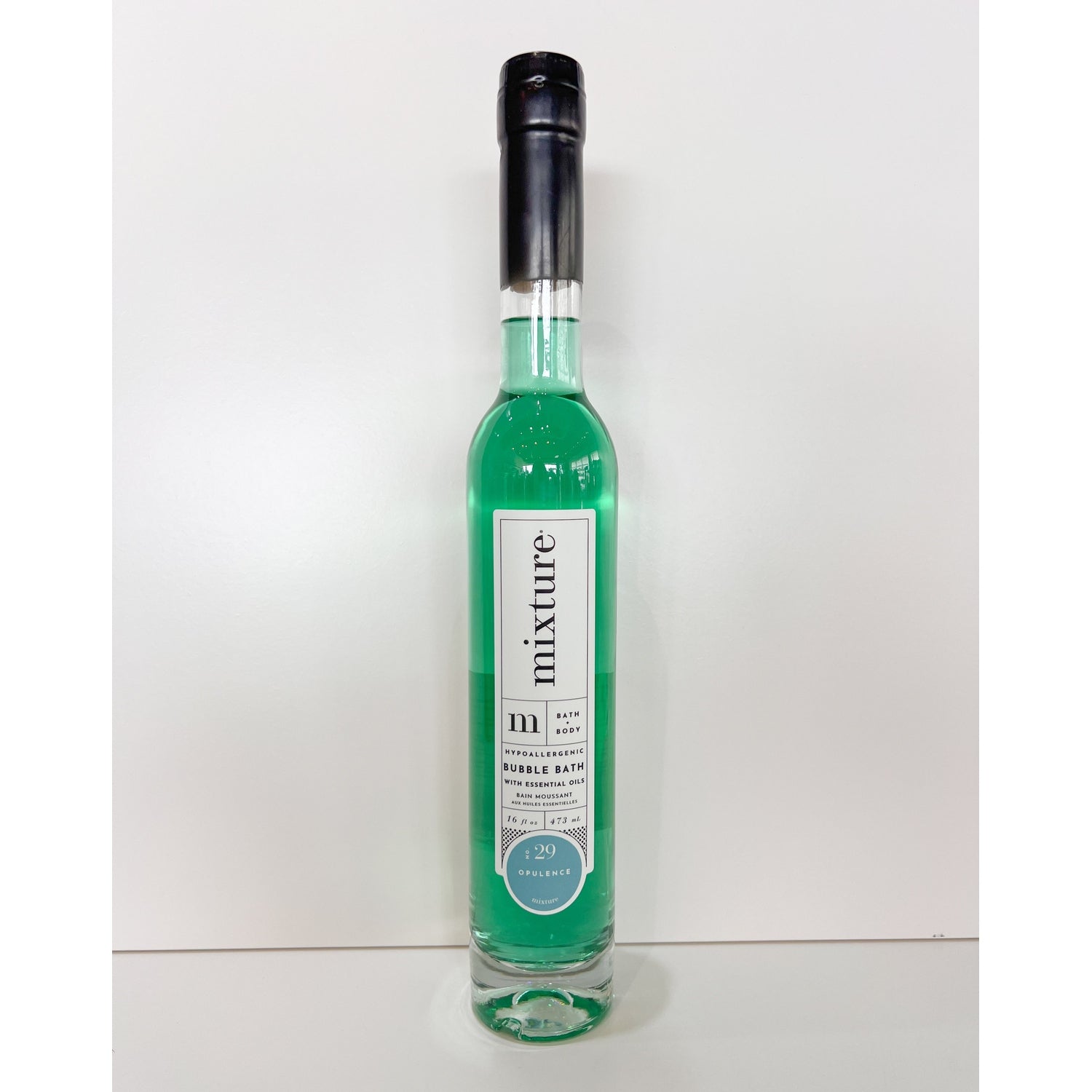 A tall slender glass bottle of Mixture bubble bath featuring vibrant aqua green liquid and a distinctive black wax-sealed top stands on a clean white surface with a minimalist white background showcasing its white label with "Mixture Bath Body" and "No 29 Opulence"
