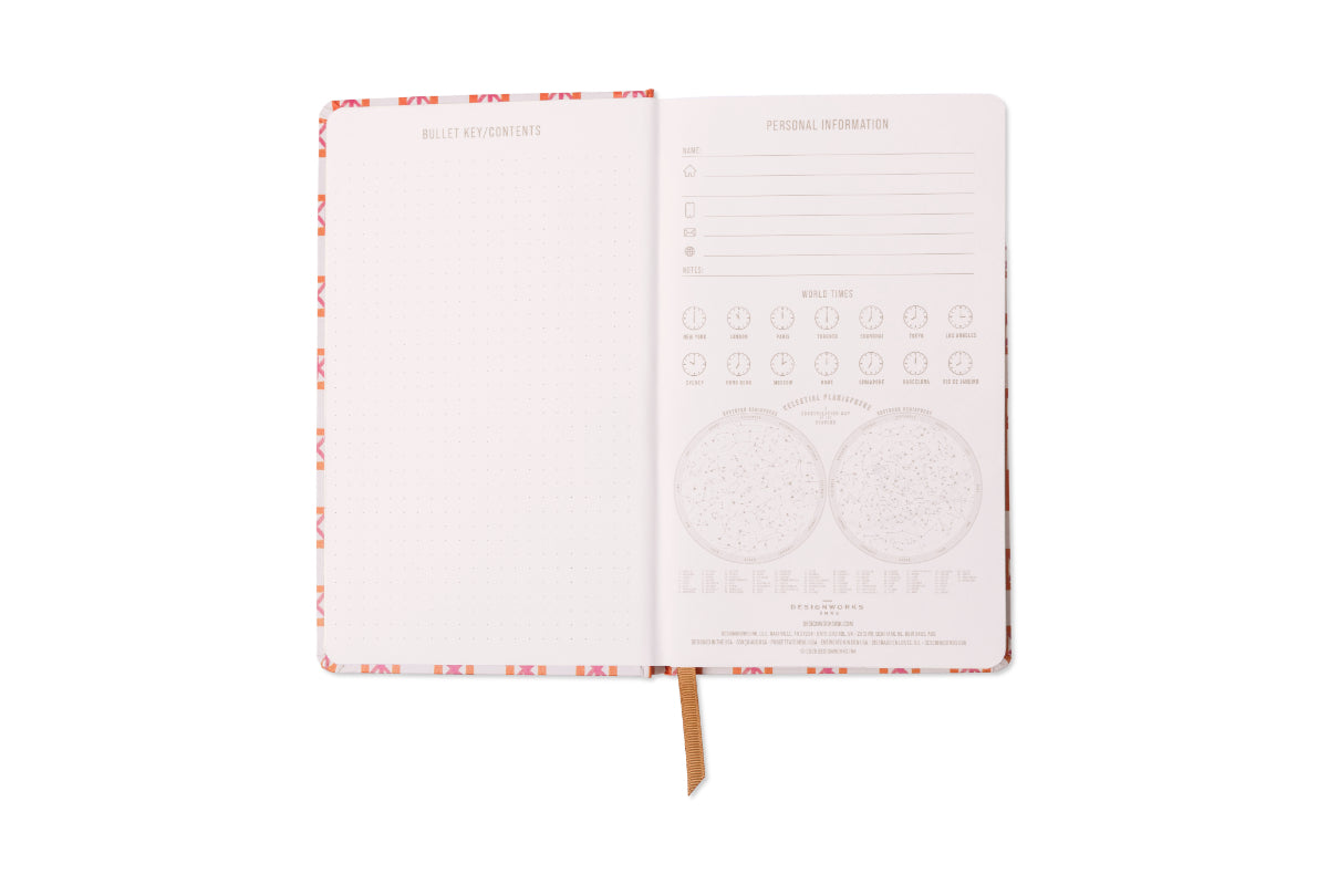 Designworks open planner displaying its orange and pink geometric patterned cover a left dot grid bullet key contents page and a right page detailing personal information world times for major cities and two celestial planisphere star charts with a brown ribbon bookmark