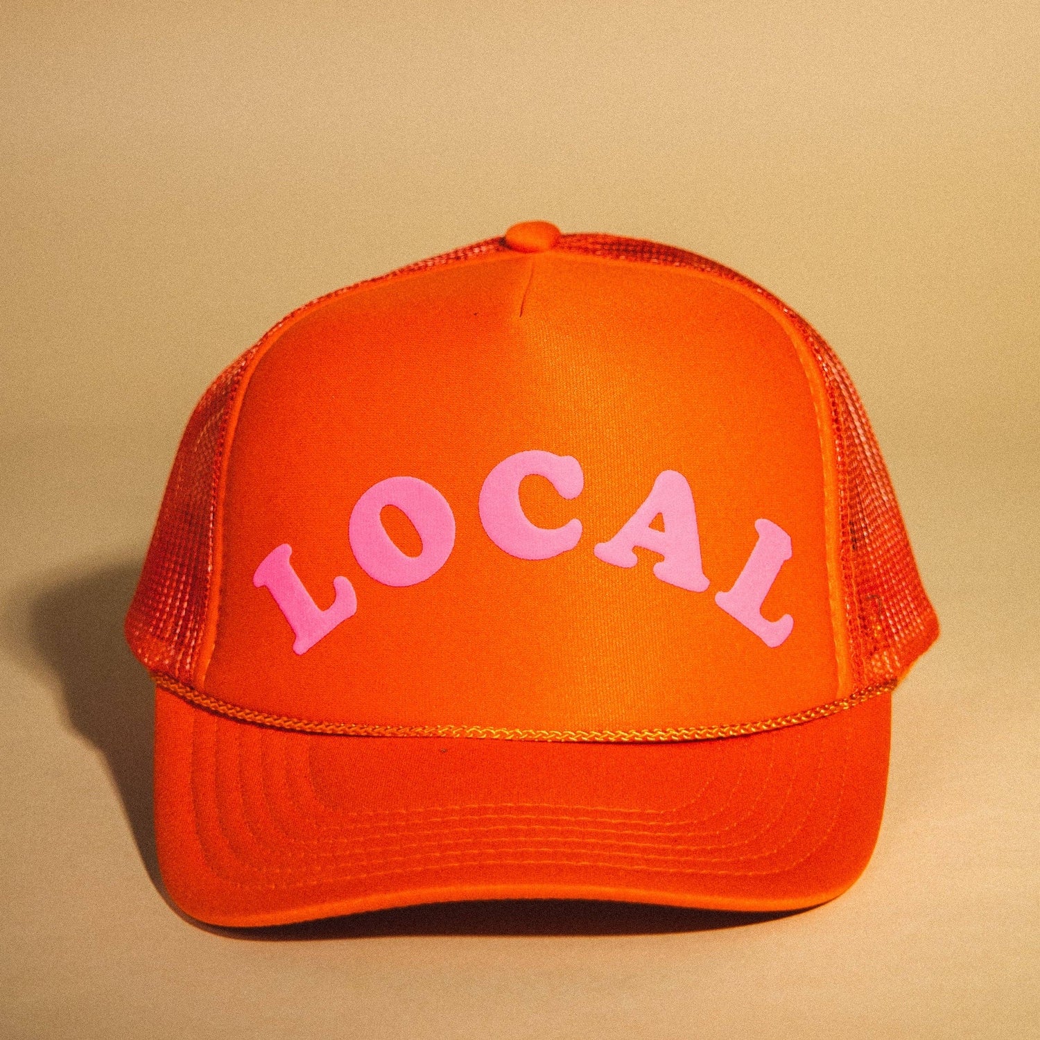 Product photograph of a bright orange trucker hat featuring the word LOCAL in prominent pink capital letters with a matching mesh back