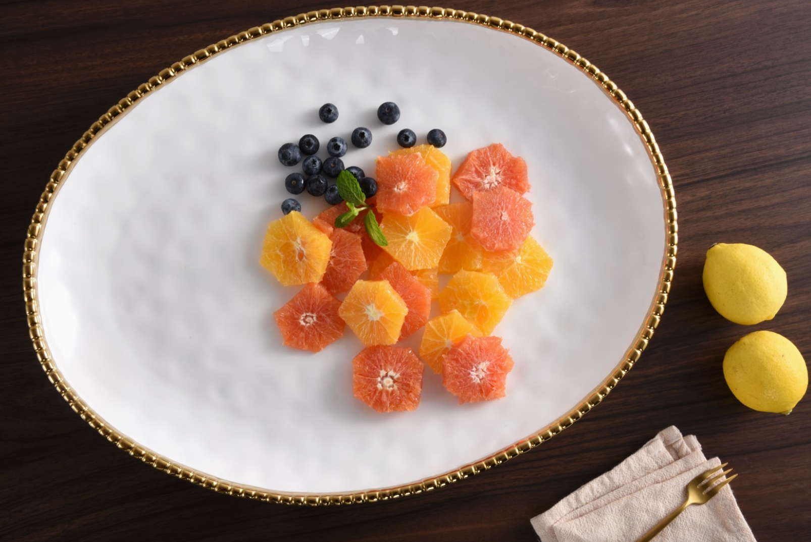 A white oval serving platter with a gold beaded rim displays hexagonally cut orange and red grapefruit segments and fresh blueberries with a mint sprig on a dark wood table beside two whole lemons and a beige napkin