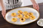 A white oval platter with a beaded gold rim holds eight Portuguese egg tarts featuring golden-brown flaky crusts and creamy yellow custard with caramelized tops, presented by a person in a KITCHEN apron, with a small white teacup beside it on a wooden surface