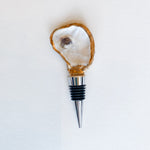 Handcrafted mixed media wine bottle stopper featuring a natural oyster shell with hand-painted gilded gold edges and a dark purple muscle scar atop a polished stainless steel base against a white textured background