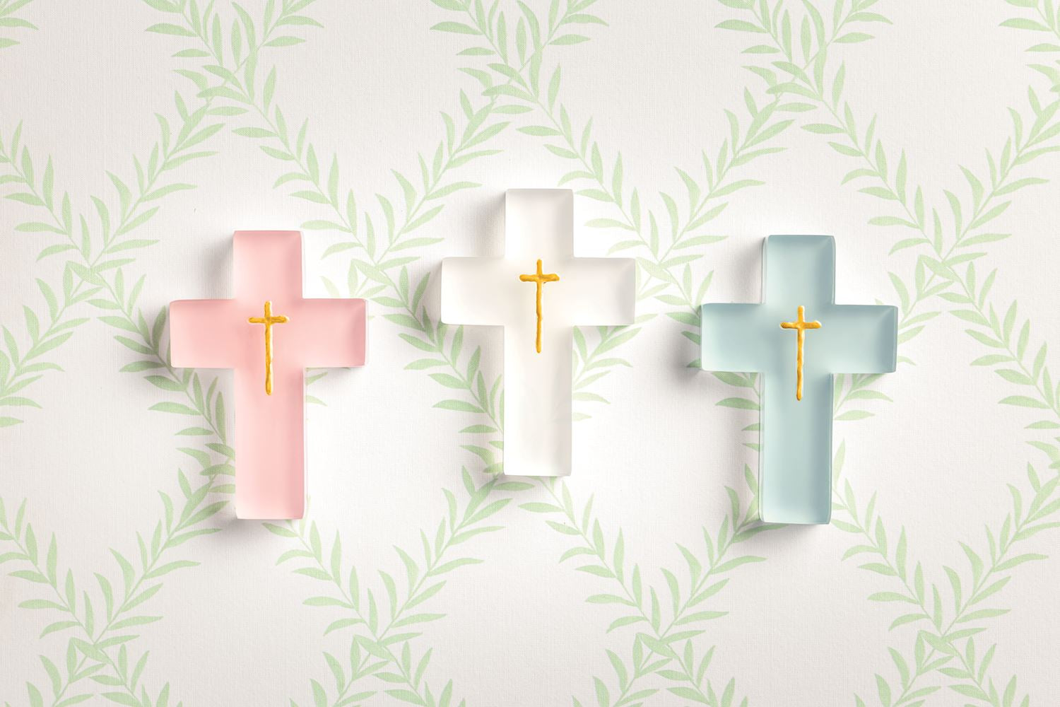 Three frosted glass crosses in pastel pink, white, and teal, each with an embossed golden cross, set against a white background with a repeating light green leaf pattern