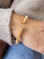 Photograph of a woman’s wrist wearing an open gold cuff bracelet with a delicate woven mesh texture and distinct ribbed cylindrical ends, styled with a beige knit sweater and blue jeans