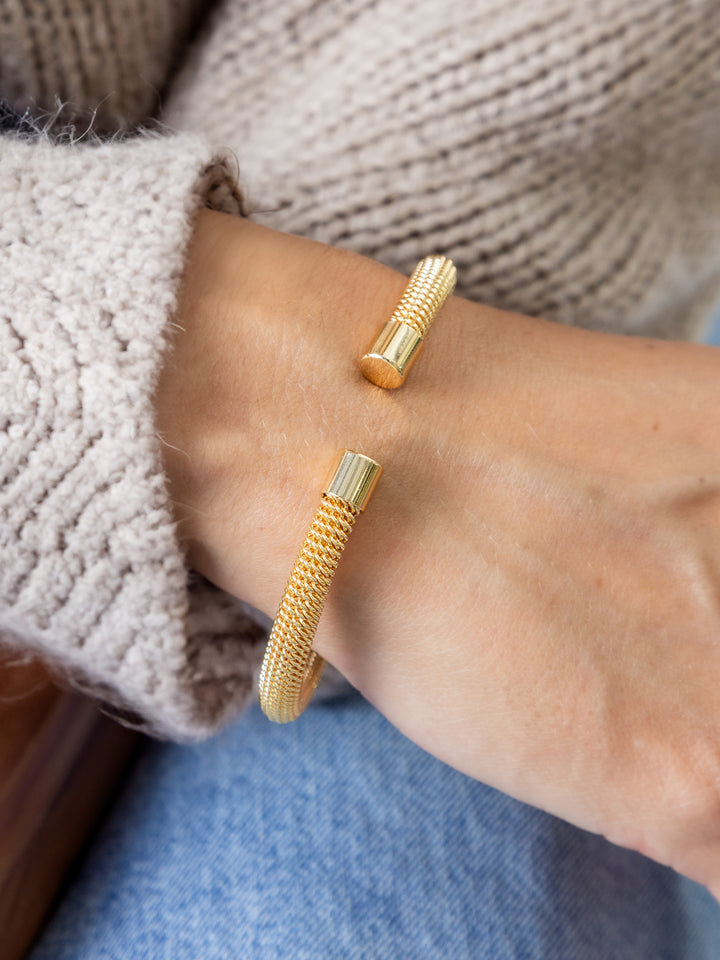Photograph of a woman’s wrist wearing an open gold cuff bracelet with a delicate woven mesh texture and distinct ribbed cylindrical ends, styled with a beige knit sweater and blue jeans