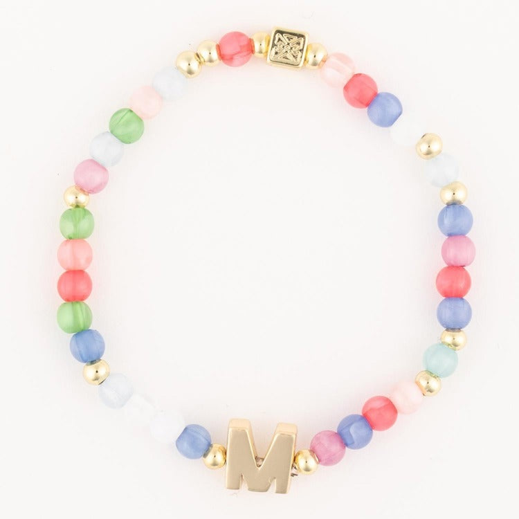 A colorful elastic bracelet featuring pastel round beads in various shades, a prominent gold initial M charm, and gold accent beads including a detailed square bead