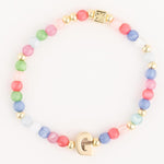 A pastel multi-colored beaded bracelet featuring round glass beads, small gold accent beads, a prominent gold G initial charm, and a square gold charm with an embossed design